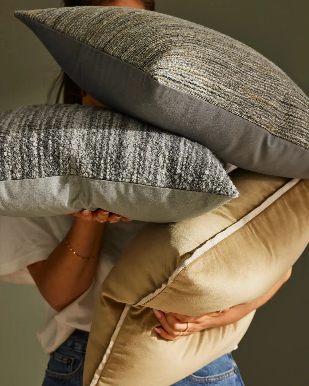 Stacked neutral cushions in grey and cream tones showcase textural linen blends and wool weaves against soft sage backdrop, styled with natural linen in warm sand hues