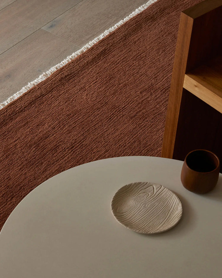 Warm terracotta wool rug with fringe edge anchors minimalist dining scene, pairing earthy tones with natural wood and cream ceramics