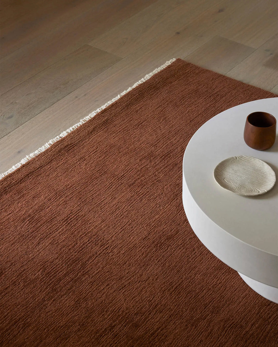 Warm terracotta wool rug anchors minimalist interior with white side table, natural wood flooring, and earthy ceramic accessories in neutral tones