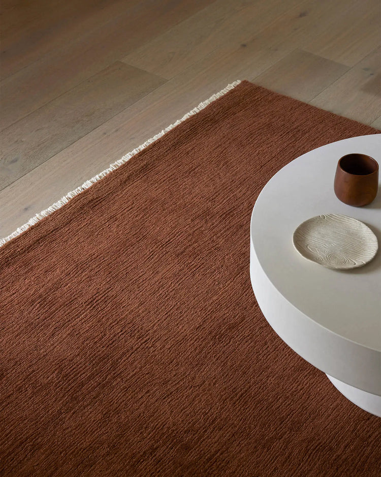 Warm terracotta wool rug anchors minimalist interior with white side table, natural wood flooring, and earthy ceramic accessories in neutral tones