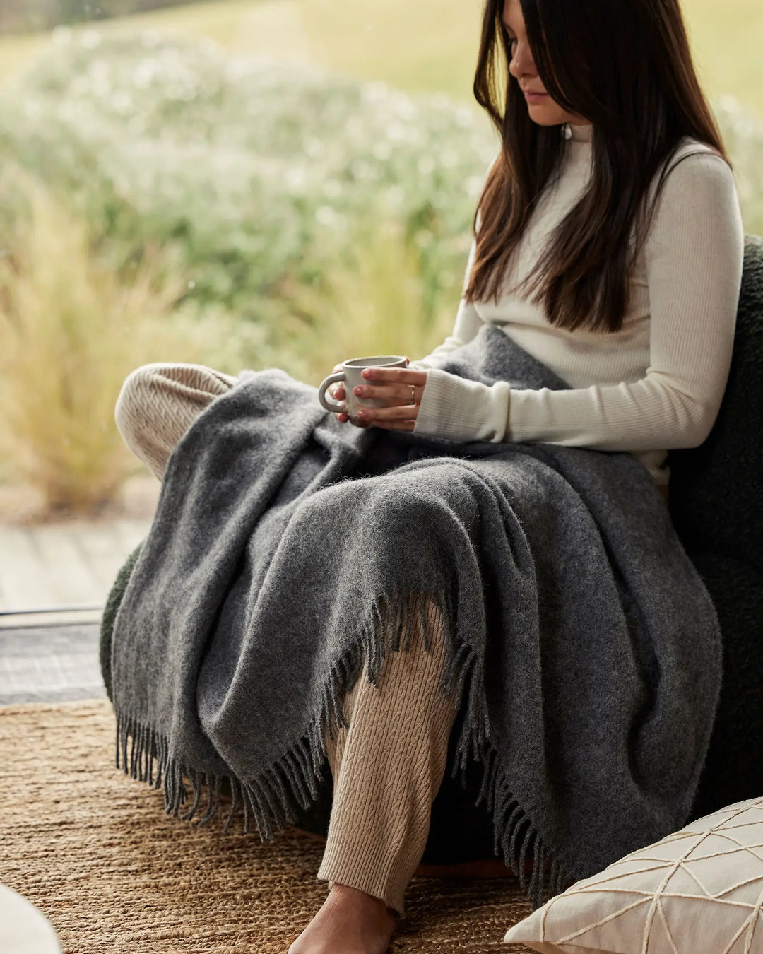 Woman seated by window in cream ribbed knit, draped in charcoal wool throw with fringed edge, holding mug with soft green landscape beyond