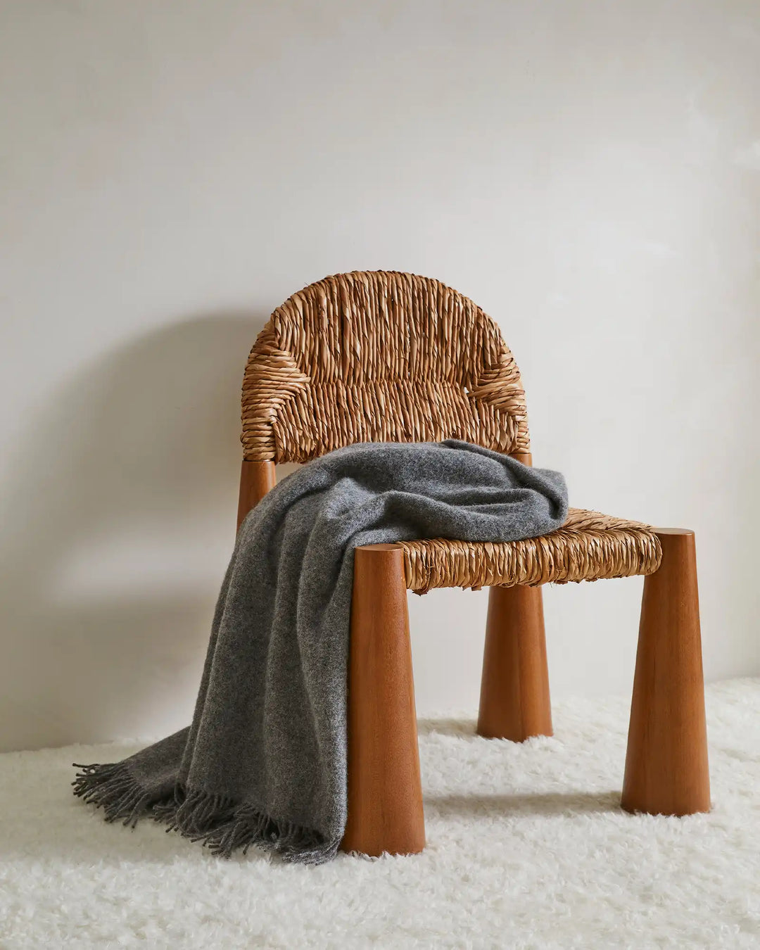 Nevis Charcoal lambswool throw in mid-grey with tassel fringe border, draped over woven chair by Weave Home