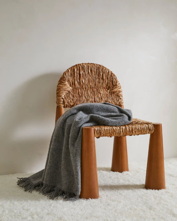 Nevis Charcoal lambswool throw in mid-grey with tassel fringe border, draped over woven chair by Weave Home