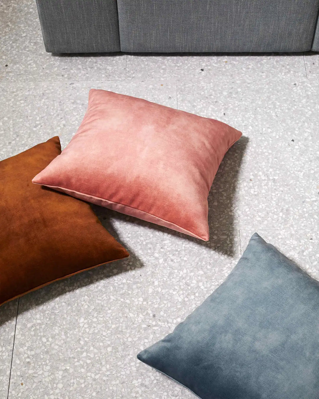 Ava cushions styled on concrete floor in warm terracotta, soft blush pink and slate blue velvet, with charcoal sofa above