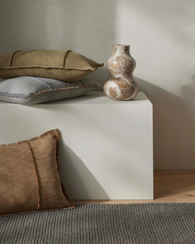 Neutral minimalist styling with stacked textiles in earthy tones, featuring Adela Cocoa cushion with frayed edges beside ceramic vessel on white cube, warm taupe and cream palette