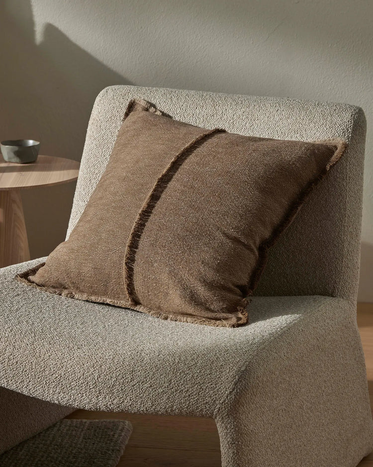 Neutral living room with cream textured armchair layered with cocoa and taupe linen cushions, natural light casting shadows on wall, minimalist aesthetic with warm earth-tone palette