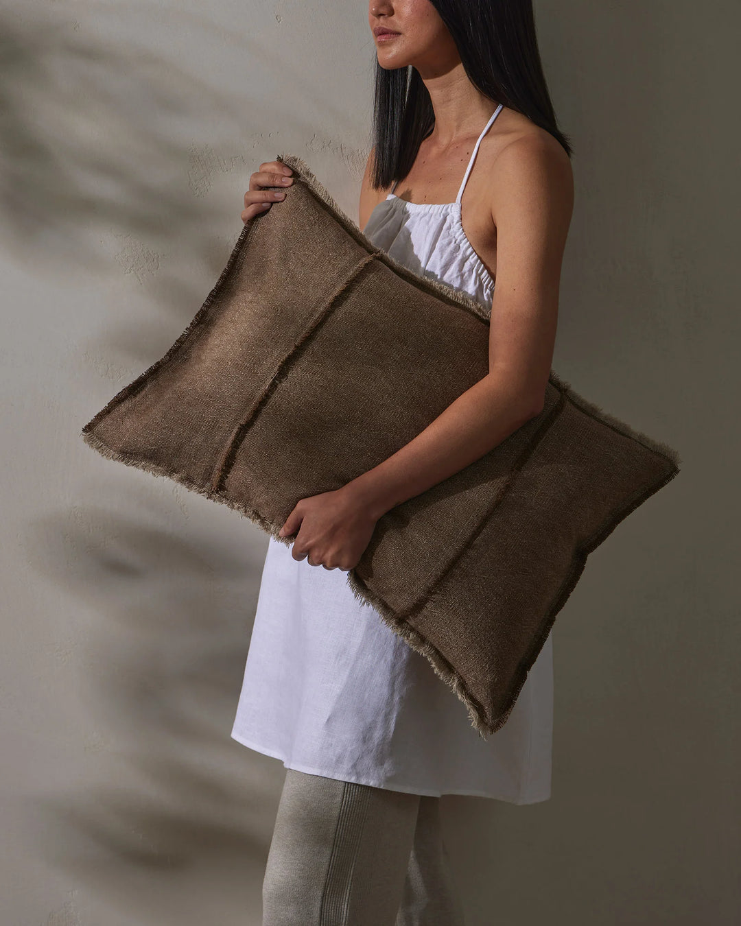 Woman holding taupe linen cushions against white textured wall, styled with white cotton dress and neutral palette, showcasing soft woven textures and earthy tones