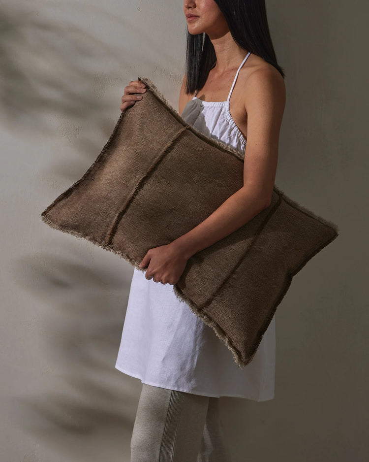 Woman holding taupe linen cushions against white textured wall, styled with white cotton dress and neutral palette, showcasing soft woven textures and earthy tones