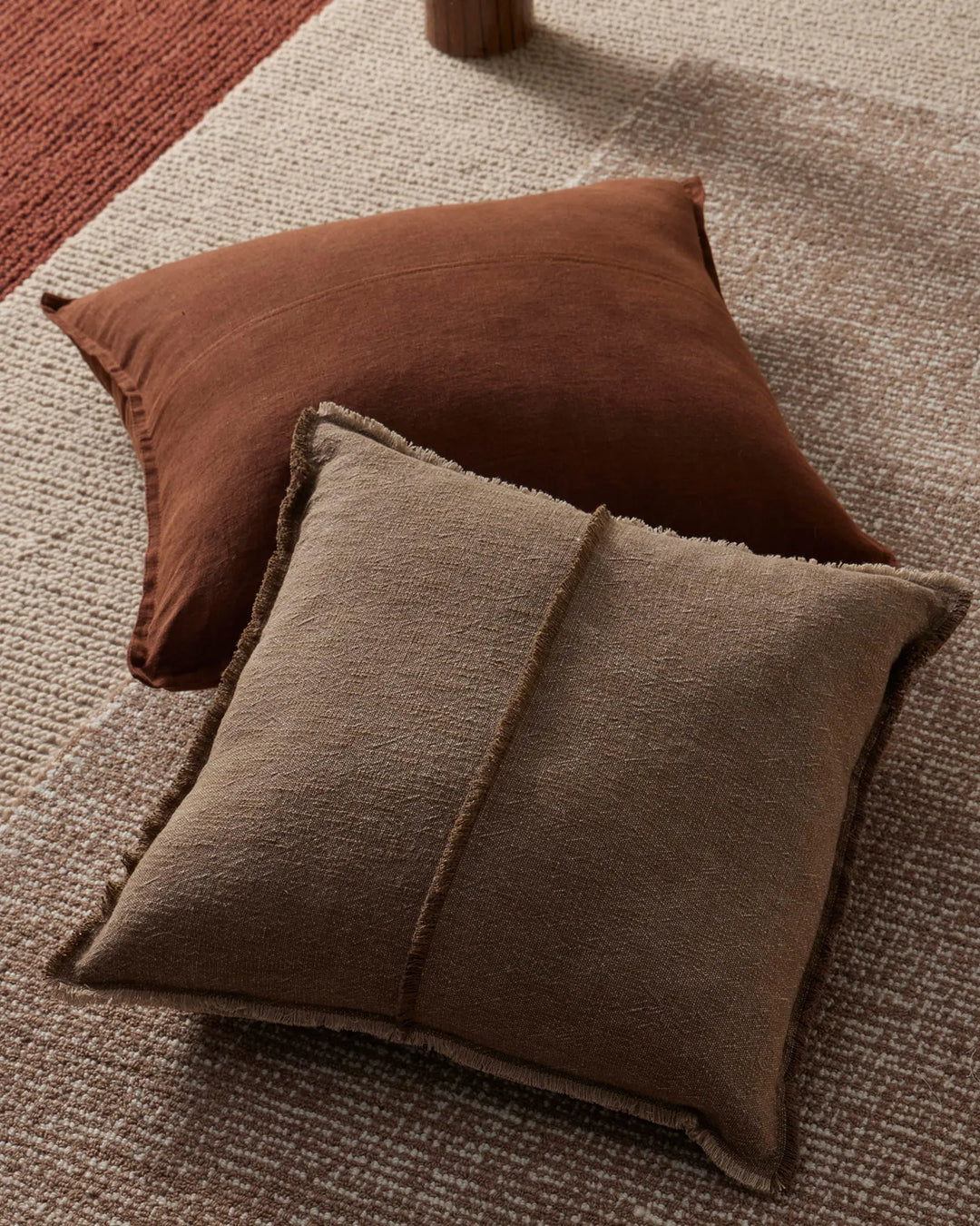 Warm earth-toned cushions in cocoa and natural linen textures layered on neutral oatmeal and terracotta woven rugs, creating a tactile, monochromatic interior styling moment