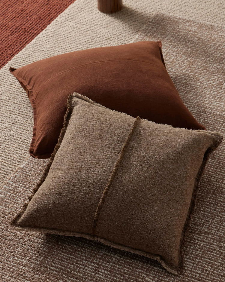 Warm earth-toned cushions in cocoa and natural linen textures layered on neutral oatmeal and terracotta woven rugs, creating a tactile, monochromatic interior styling moment