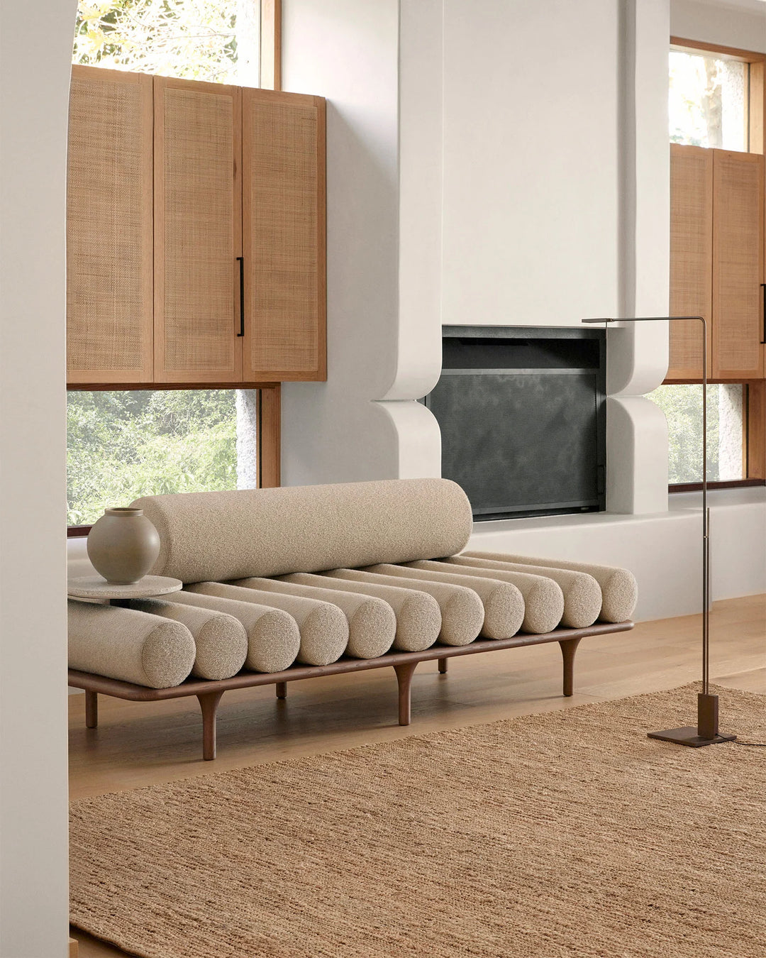 Mid-century modern living room with natural jute rug, beige linen daybed with cylindrical bolsters, rattan cabinetry, and minimalist timber frame—earthy, neutral palette with warm natural light