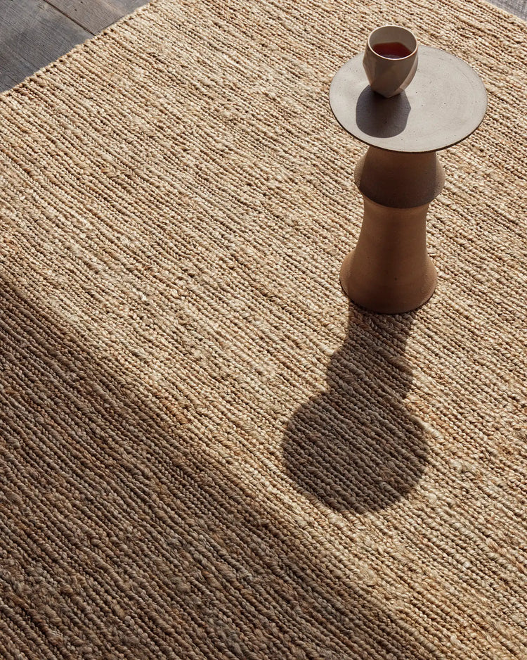 Neutral jute rug with dense woven texture in warm natural tones, styled with minimalist side table and ceramic cup casting soft shadows