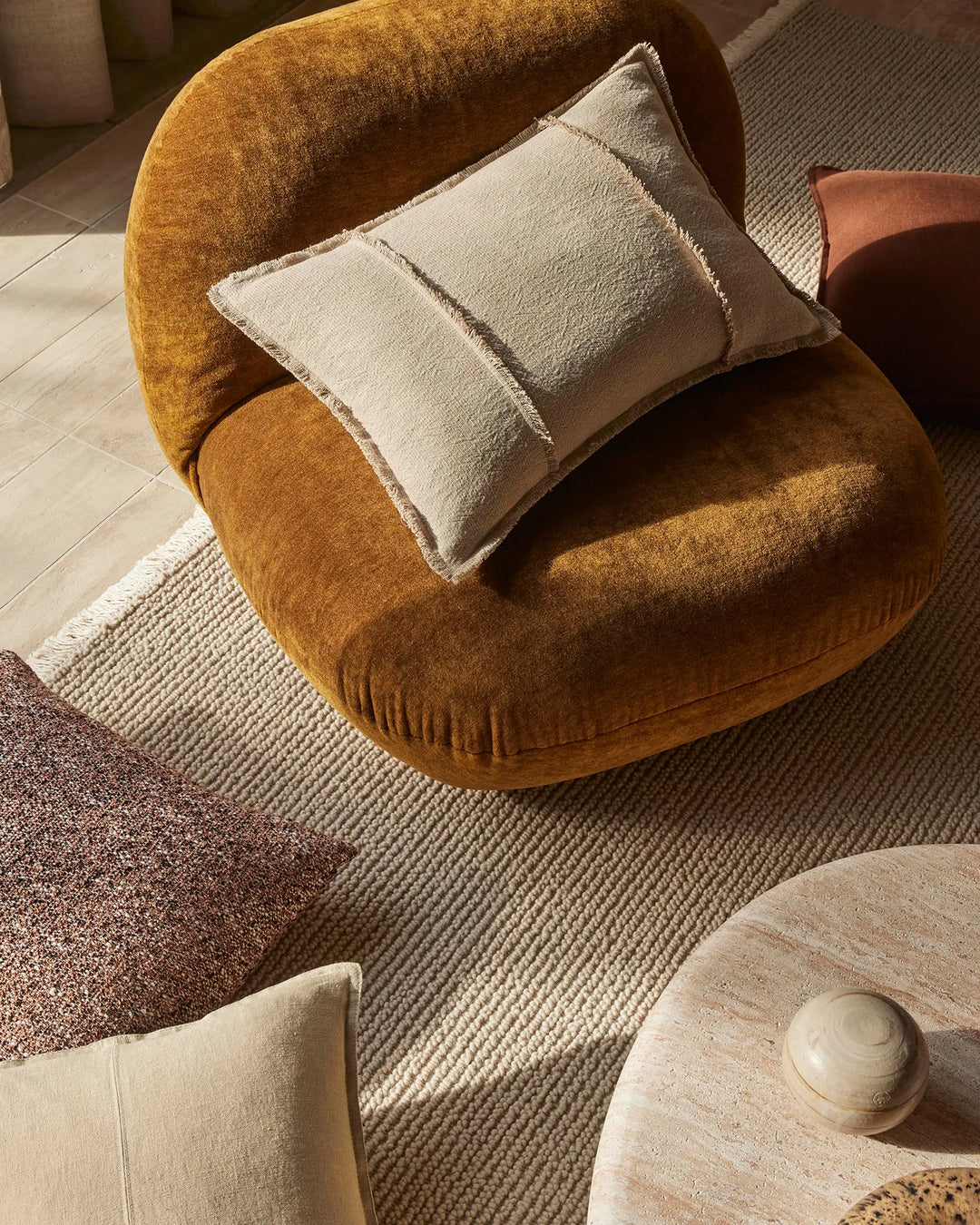 Warm ochre rounded chair styled with cream linen cushions on textured stone rug, earthy modern interior with natural wood and soft furnishings