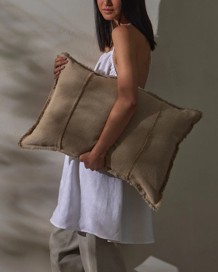 Woman in white linen dress holding oversized natural linen cushion with raw frayed edges against textured white wall, minimalist neutral aesthetic