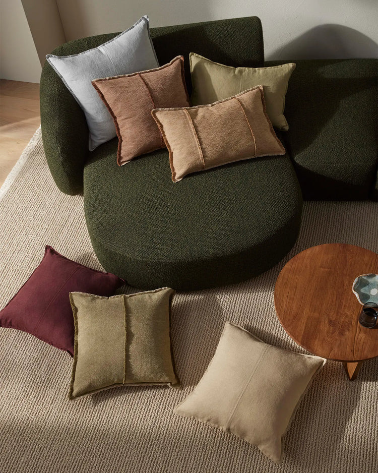 Earthy-toned Adela cushions layered on a forest green curved armchair with warm terracotta, sage and cream accents against natural linen flooring, minimalist styling mood