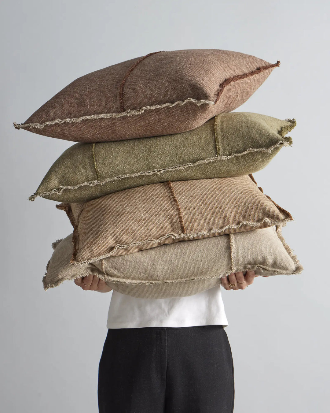 Stacked Adela cushions in warm earth tones—chocolate brown, sage green, and taupe—showcasing raw-edge linen and cotton blend textures against a neutral backdrop
