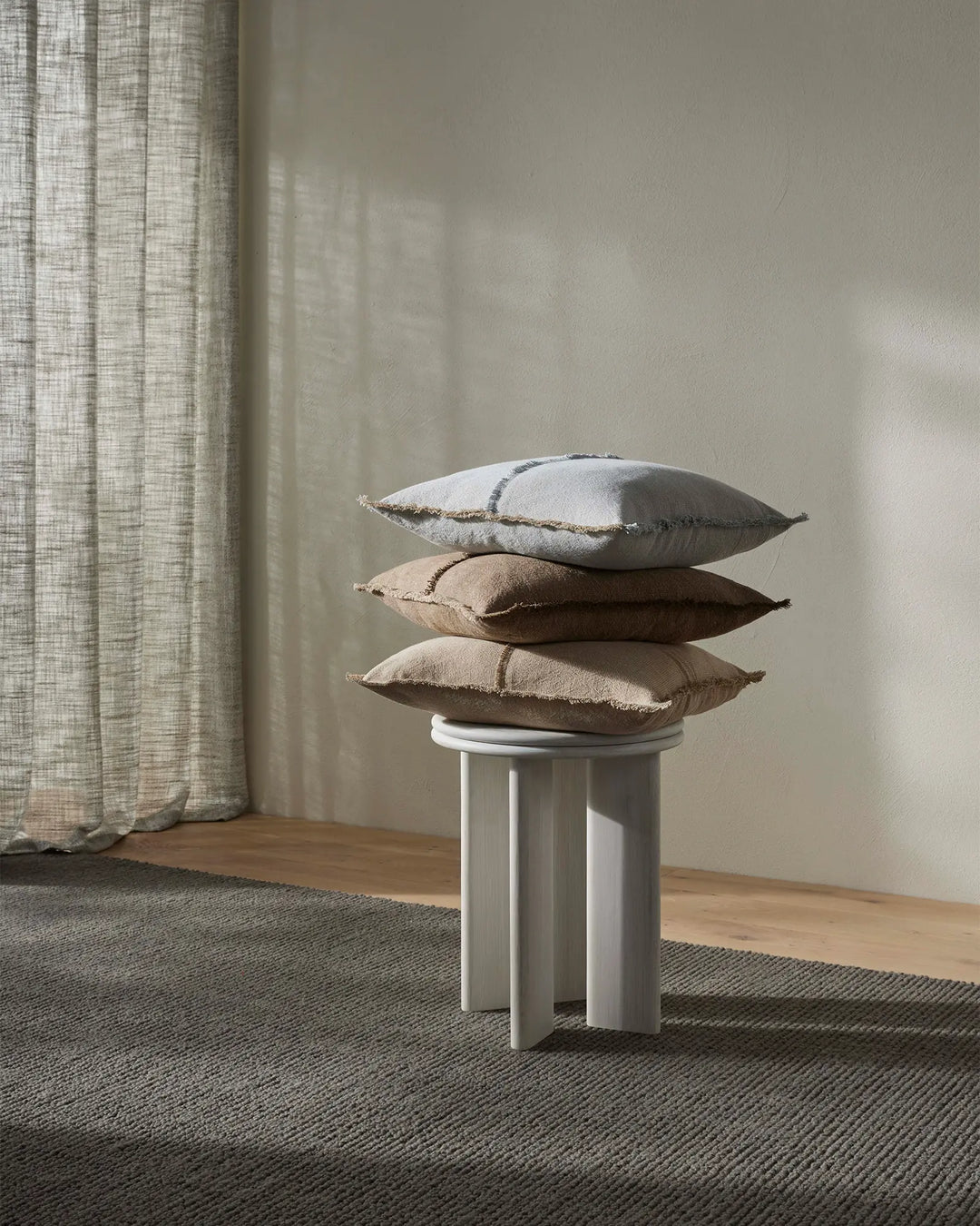 Stacked linen cushions in soft neutrals—haze, cashmere, and cocoa—on a white pedestal amid natural light, textured rug, and oatmeal linen curtains