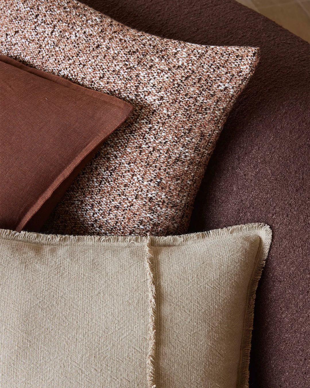 Layered terracotta, cream and chocolate cushions in textured linen and linen blends arranged on warm-toned upholstery, showcasing earthy tones and tactile fabric contrasts