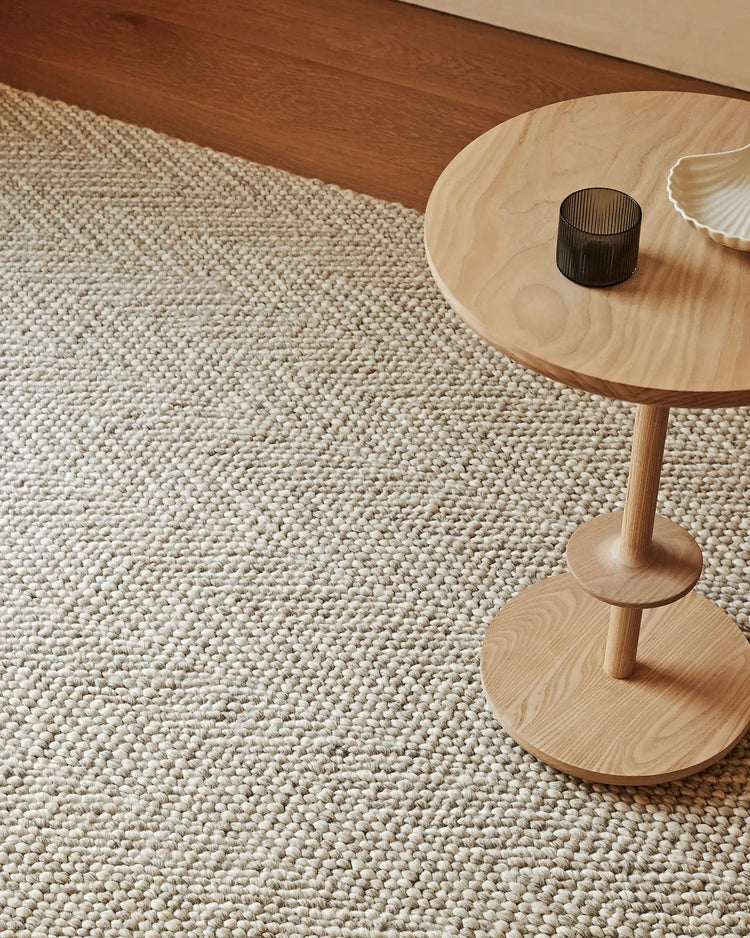 Neutral minimalist bedroom with Zambesi Sandstorm wool-blend rug in warm beige, timber side table, and pale wood flooring creating a cohesive earthy palette