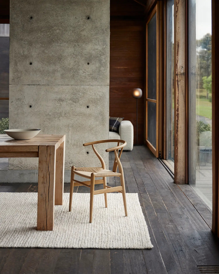 Minimalist dining space with concrete wall, timber table, wooden chair, and neutral Suffolk Pearl jute rug anchoring the serene earth-tone palette