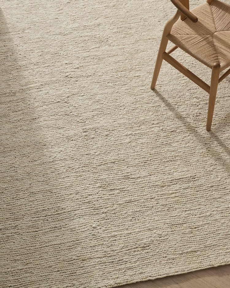 Minimalist Scandinavian interior with natural jute texture rug in cream, woven chair with rush seat, and warm timber legs casting soft shadows on neutral flooring.