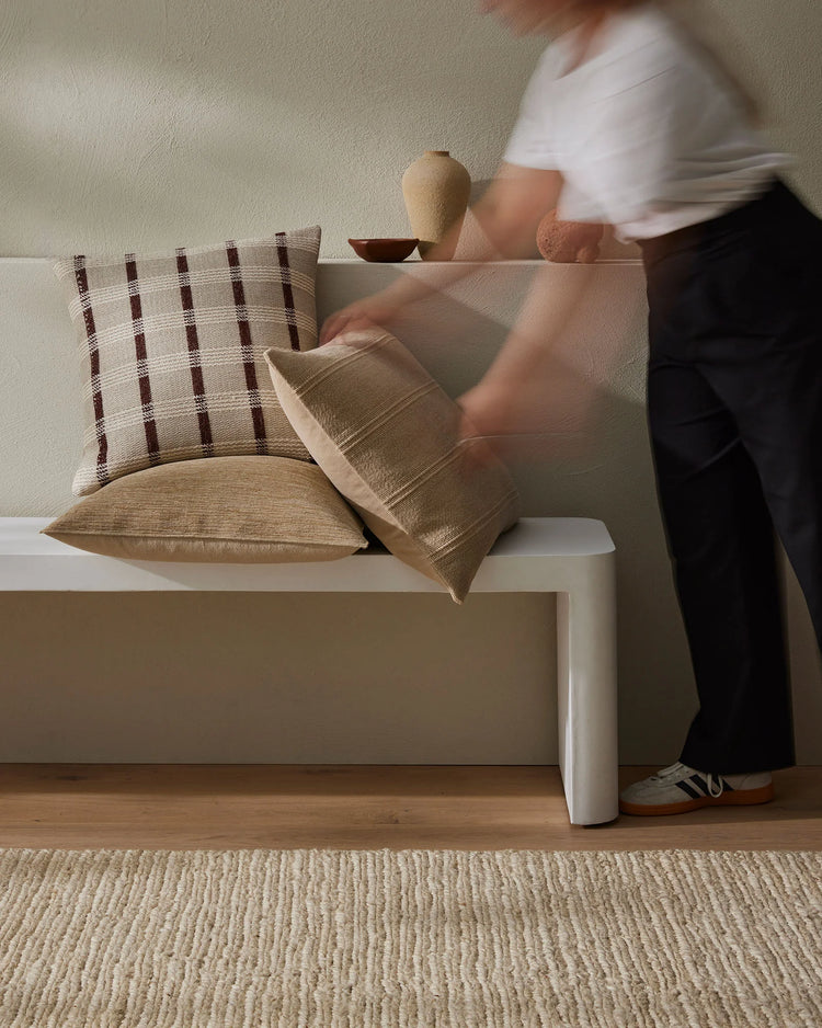 Neutral minimalist bedroom styling with warm earth-tone cushions layered on white bench, featuring check-pattern and solid textures in linen and polyester, natural timber flooring, and tonal aesthetic