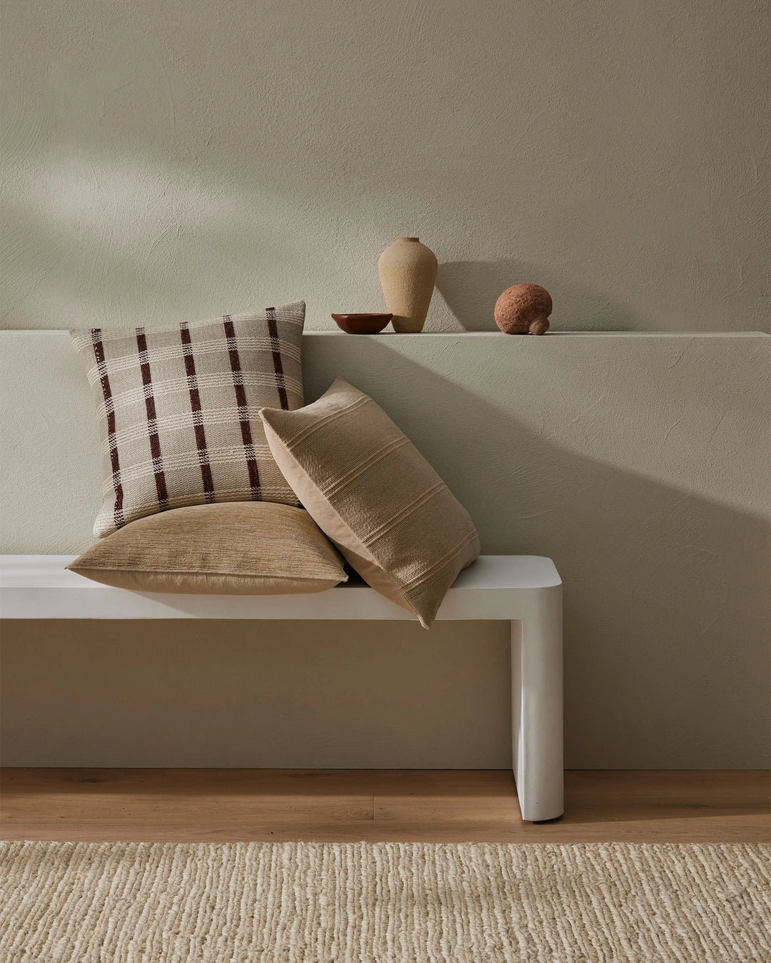 Neutral minimalist bedroom setting with layered throw pillows in cream and rust tones featuring geometric grid pattern and textured weave, styled on white platform bed with natural wood flooring and woven rug
