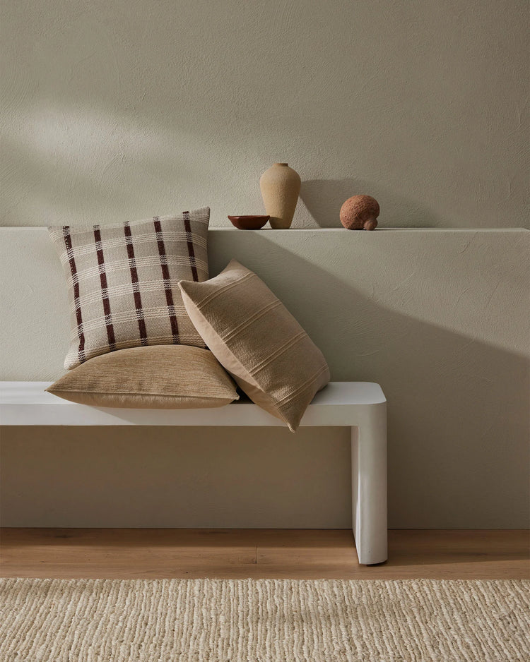 Neutral minimalist bedroom setting with layered throw pillows in cream and rust tones featuring geometric grid pattern and textured weave, styled on white platform bed with natural wood flooring and woven rug