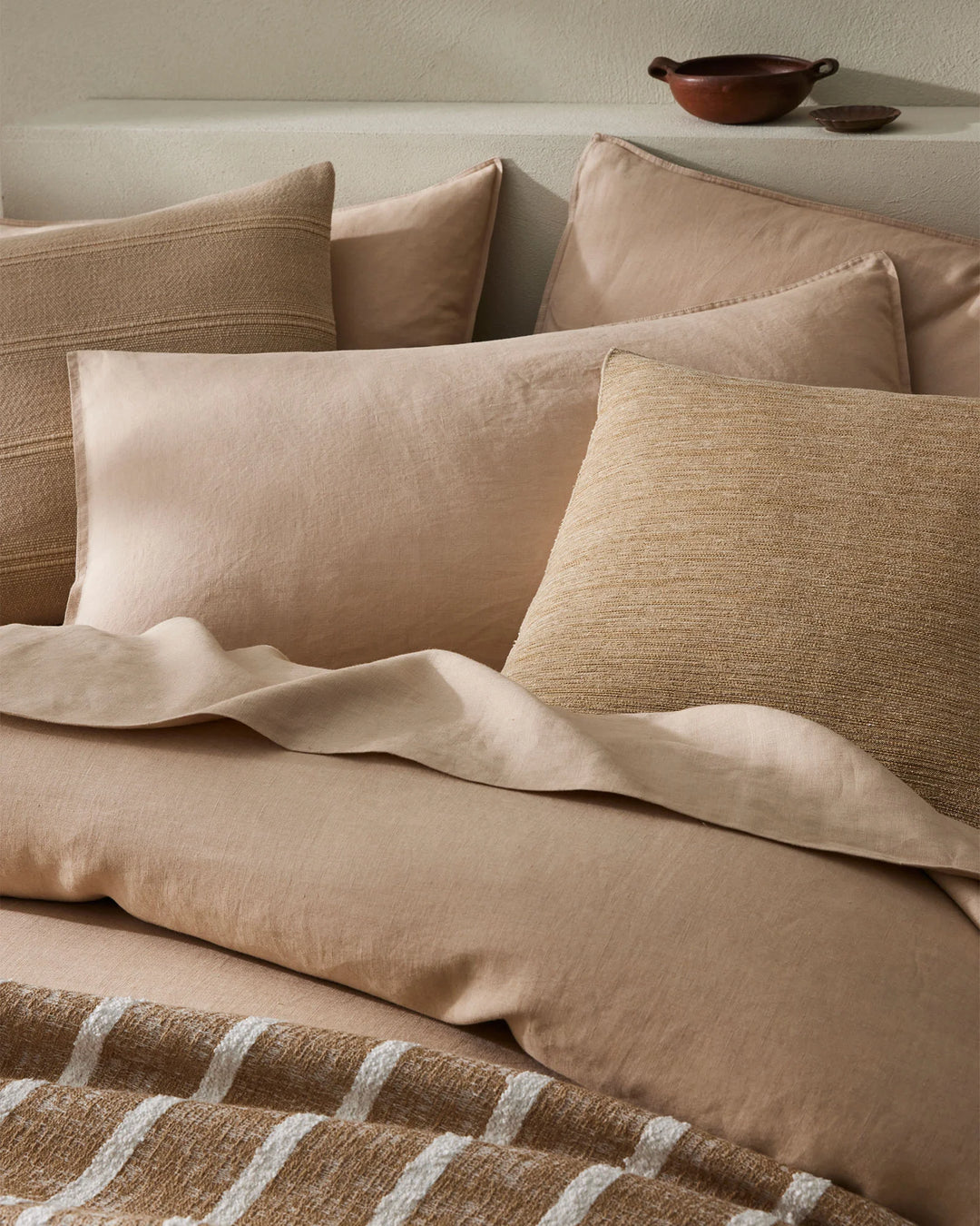 Warm neutral bedding styled with textured cushions in cream and natural tones, striped throw, and terracotta vessels on cream wall backdrop