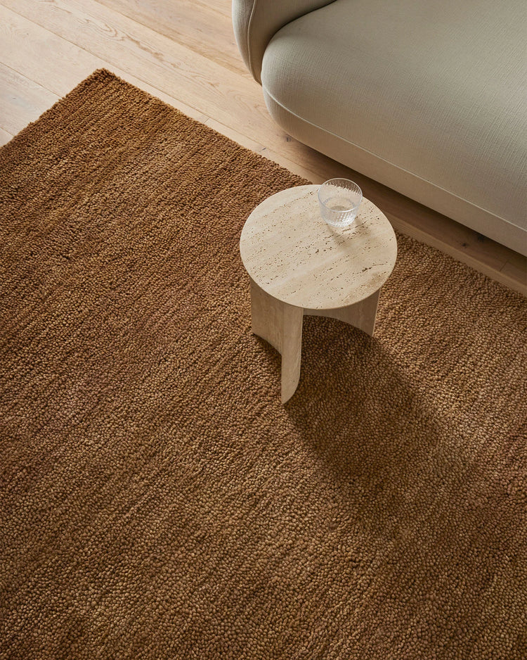Warm toffee wool rug anchors a minimalist living room with neutral upholstered chair, light oak side table, and blonde wood flooring in earthy, textural aesthetic
