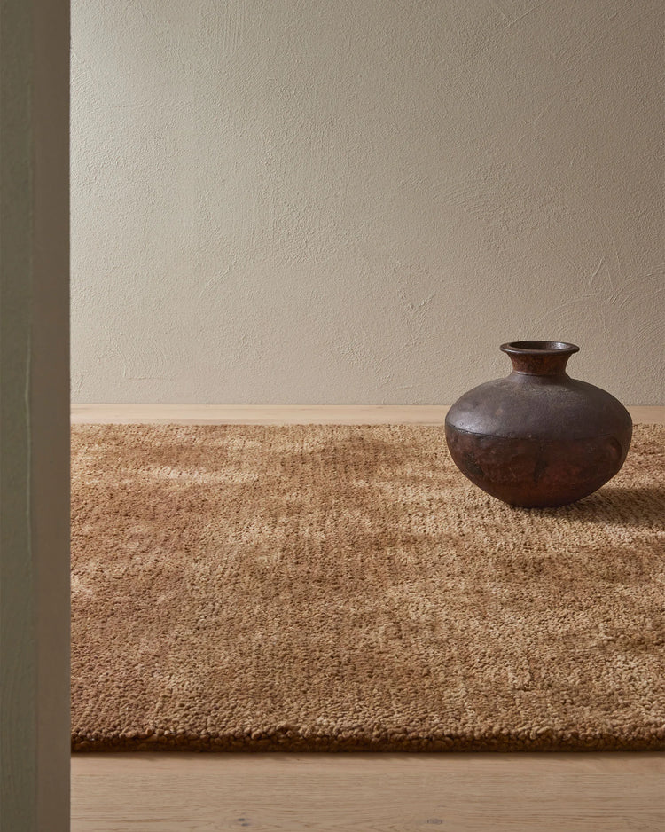 Neutral minimalist room with textured natural wool rug in toffee tone, cream plaster walls, and dark pottery vessel on wooden floor