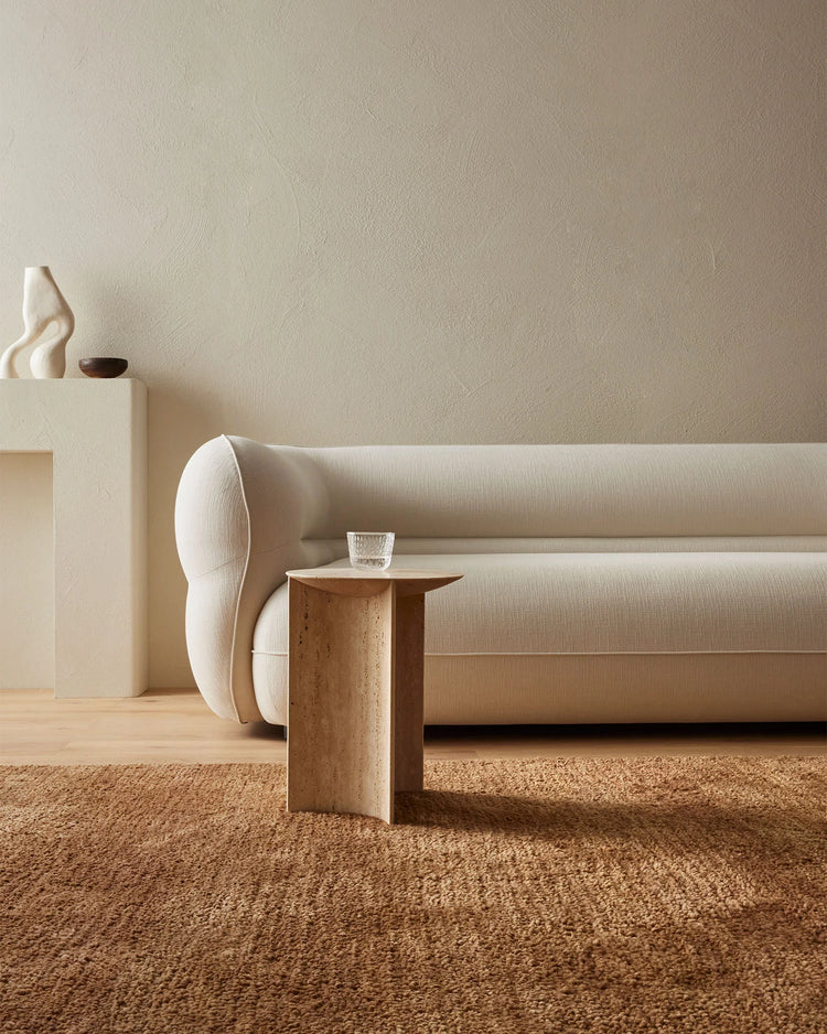 Neutral minimalist living room with cream upholstered sofa, cream pillow, wooden side table, and warm toffee-toned wool rug layering earthy textures