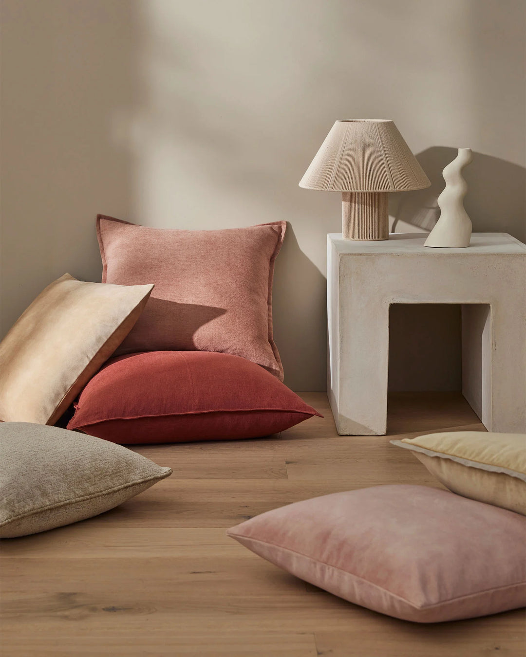 Warm earth-tone cushions in blush, burgundy, oatmeal and clay layered on wooden floor, styled with concrete plinth and textured lamp in minimalist Nordic interior