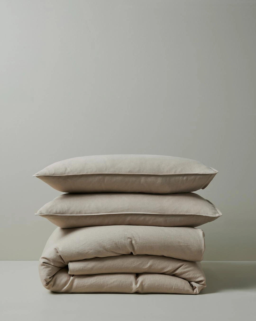 Stacked neutral bedding in soft shell tones: Ravello pillowcases and quilt cover in European linen, showcasing relaxed textures and minimalist styling against a pale grey background