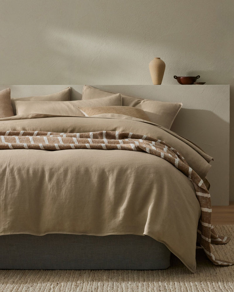 Minimalist bedroom with neutral linen bedding, layered cream and taupe tones, natural geometric patterned throw across bed, soft shadows on white headboard, warm wood accents