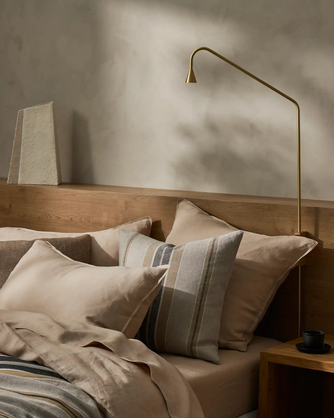 Neutral bedroom with wooden bed frame, layered linen bedding in cream and taupe tones, striped cushion, soft natural light casting shadows on pale walls