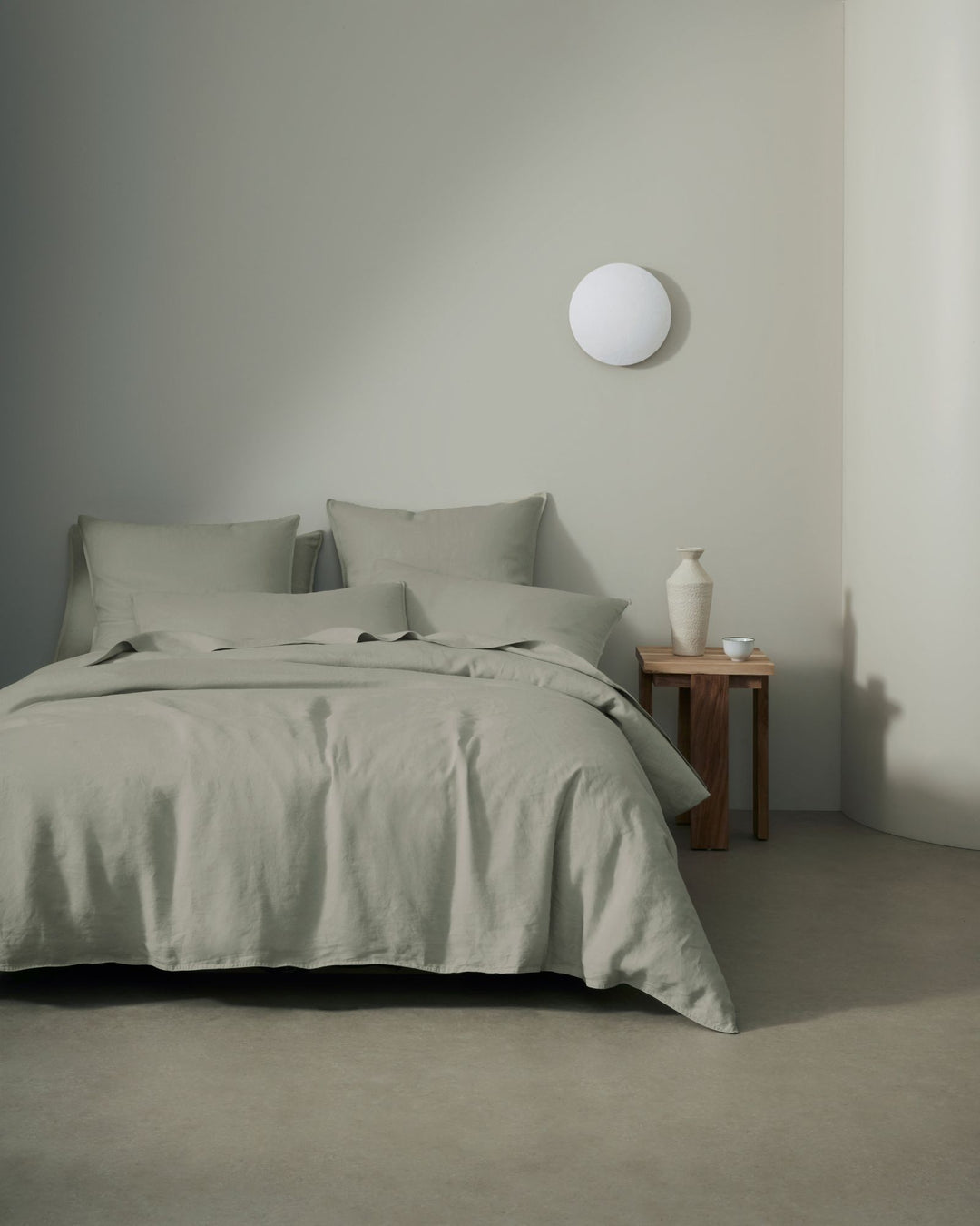 Minimalist bedroom with sage linen bedding layered on a bed, featuring European linen Ravello sheets and quilt cover in muted green tones against neutral white walls and warm wood nightstand