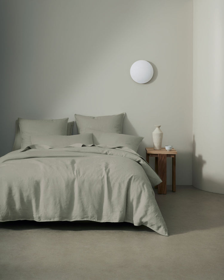Minimalist bedroom with sage linen bedding layered on a bed, featuring European linen Ravello sheets and quilt cover in muted green tones against neutral white walls and warm wood nightstand