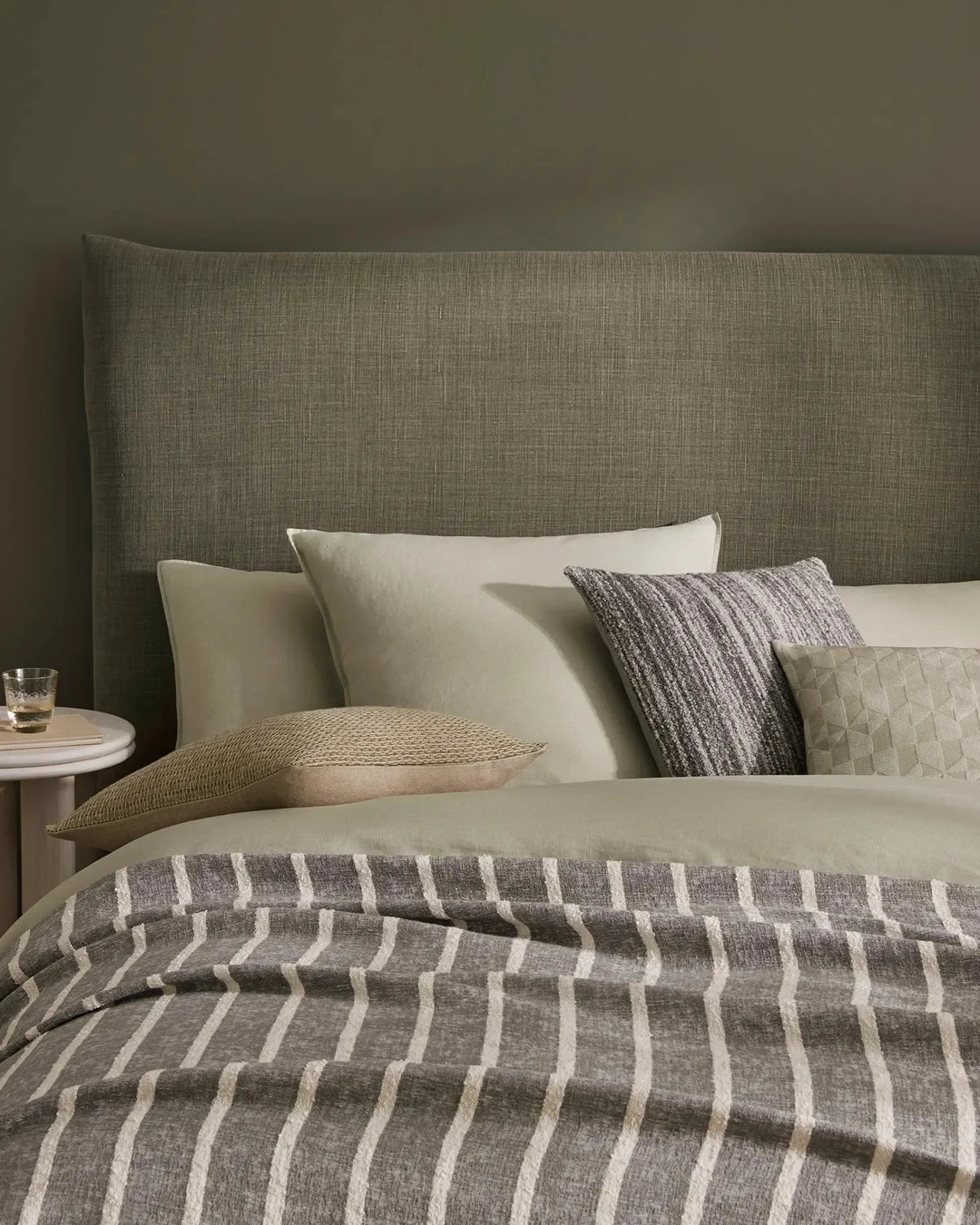 Sage linen bedding with vertical stripe duvet, layered neutral pillows in cream and charcoal textures, and upholstered headboard in earthy bedroom setting