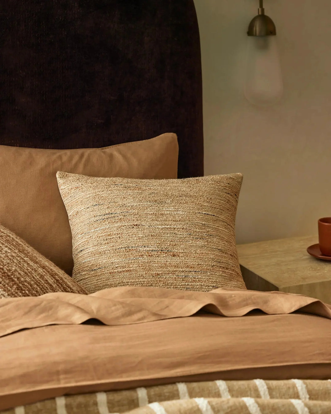 Warm neutral bedroom with layered textures: natural linen-blend cushion, caramel cotton bedding, woven throw, dark velvet headboard and brass pendant light