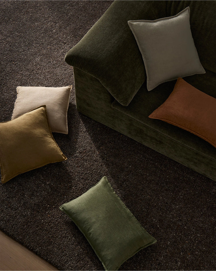 Styled cushion collection in earthy tones—olive, ochre, ecru and sage—arranged on charcoal wool rug with forest-green velvet sofa. Neutral linen-blend palette with warm, organic aesthetic.
