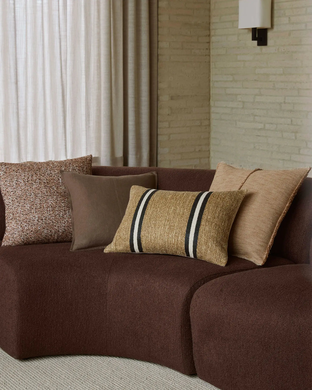 Warm earth-tone living room with chocolate curved sofa layered with textured cushions in gold, taupe and brown tones, styled with striped accent pillow and cream linen curtains