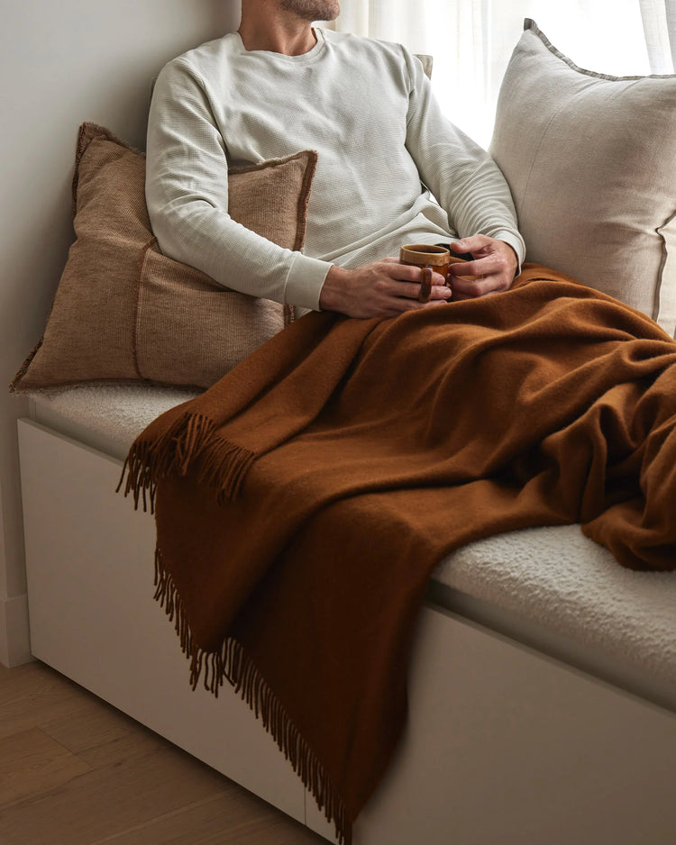 Warm earth-toned living room scene: man on cream sofa with terracotta throw and camel linen cushions, layered neutral palette with textured wool and cotton blends