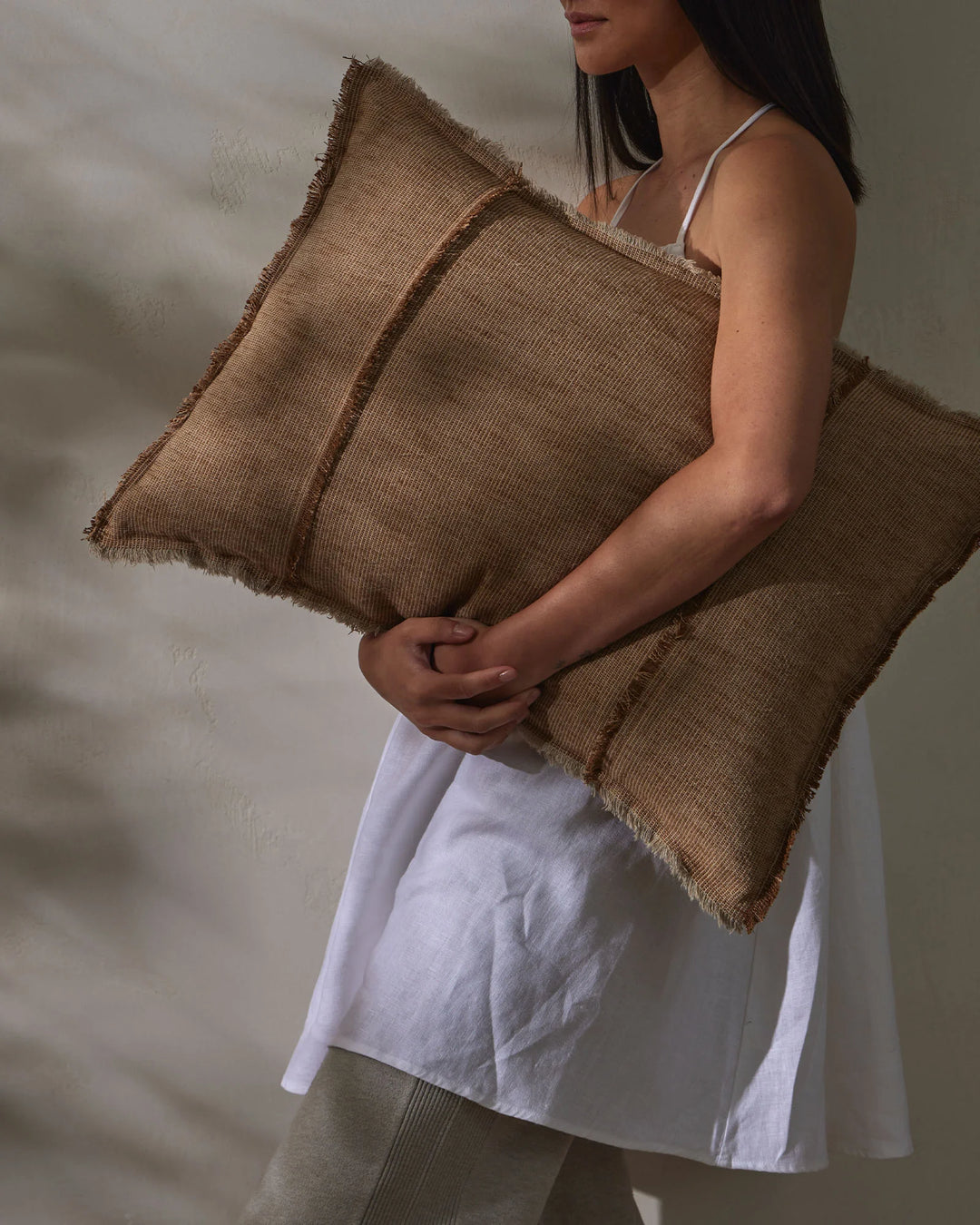 Woman holding layered taupe linen cushions against neutral backdrop, showcasing ribbed texture and raw-edge fringe detail in warm earth tones