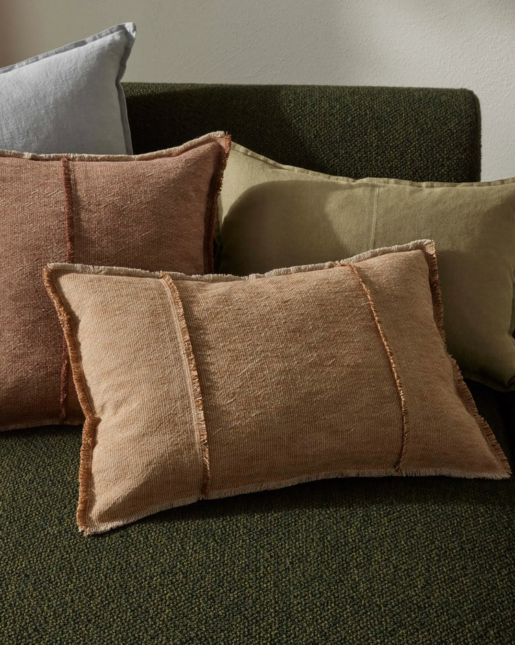 Earthy palette of layered cushions in terracotta, stone, and sage green on a forest-hued upholstered sofa, showcasing textured woven fabrics and fringed edges