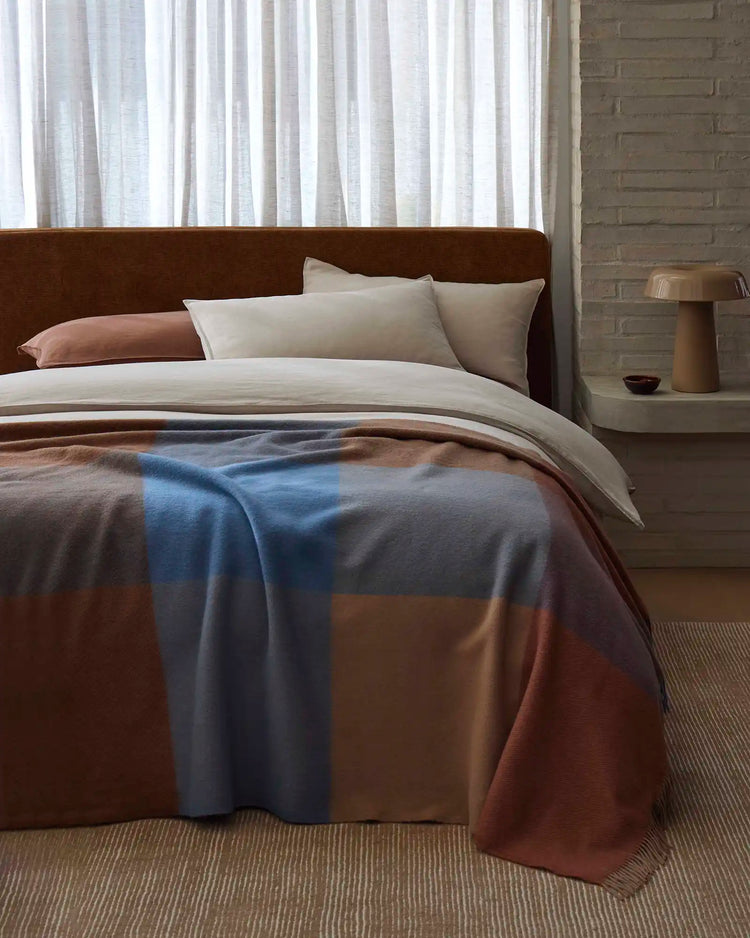 Bedroom styled with cream linens and earth-toned wool throw in blue and caramel tones across chocolate headboard, natural light filtering through sheer curtains, neutral palette with textured rug.
