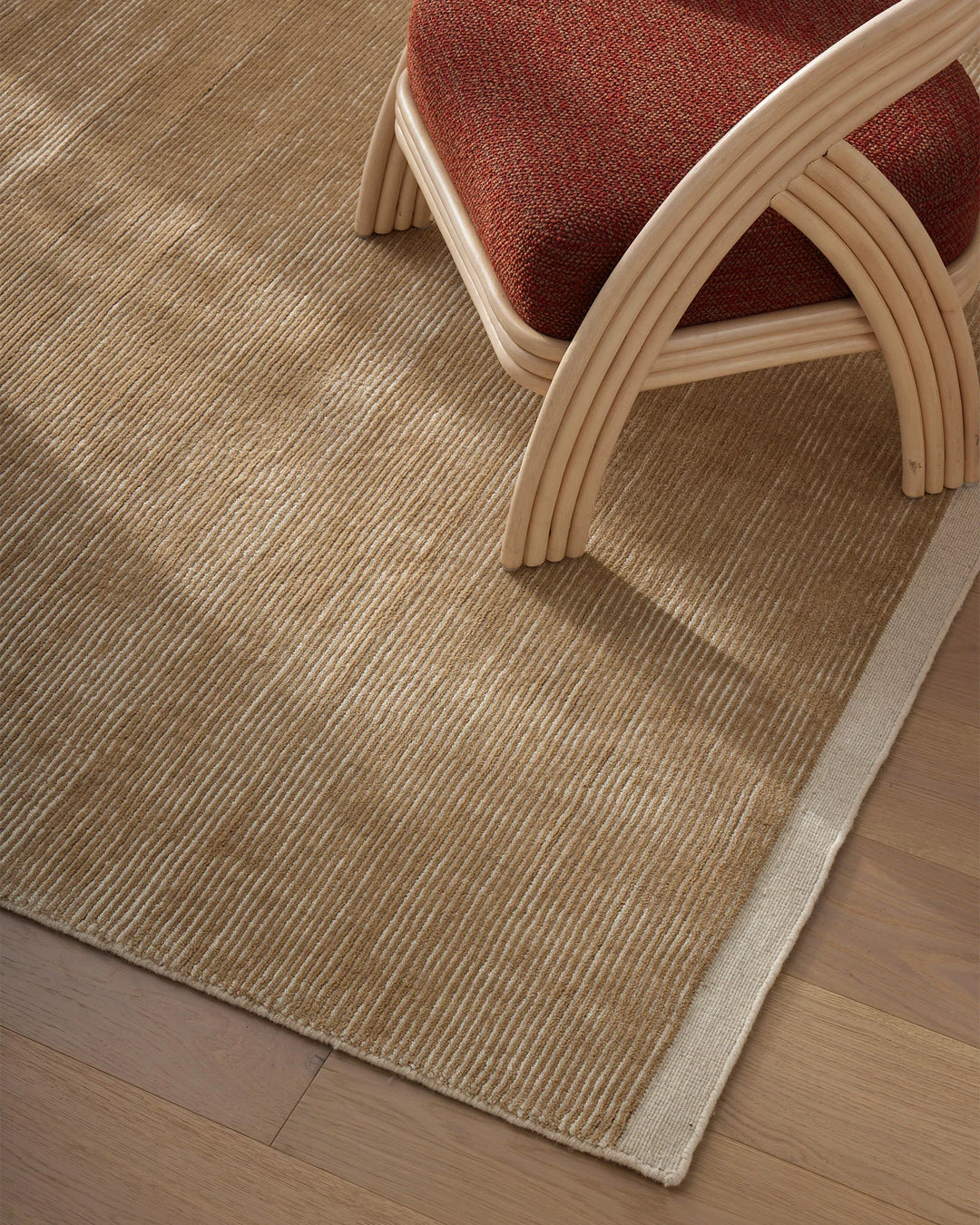 Neutral wool rug with ribbed texture anchors warm minimalist space, pairing natural beige tones with burgundy accent cushion and curved blonde wood chair frame