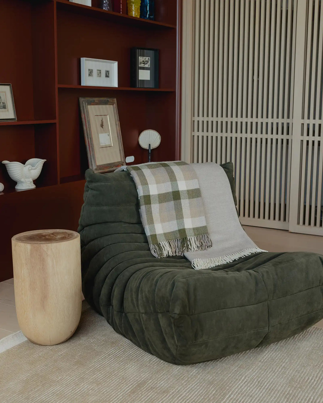 Earthy mid-century interior with sage green quilted chair, sage and cream checked wool throw, natural wood side table, and terracotta shelving unit styled with framed art and ceramic vessels.