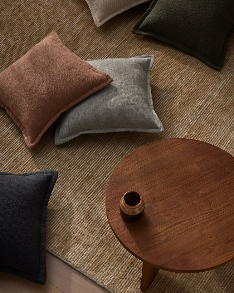 Warm, earthy interior styling with Figlio cushions in terra, kale, and mineral tones arranged on a textured wool rug beside a wooden round side table