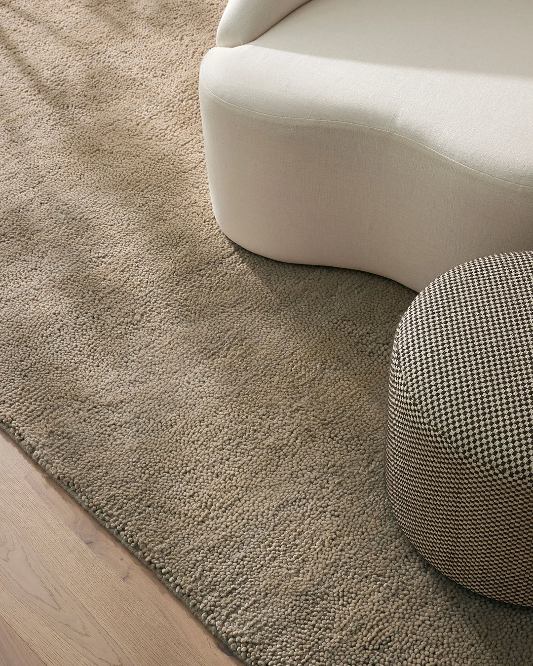 Neutral minimalist living room with cream curved furniture on a textured beige wool rug, layered with a patterned pouf, above pale timber flooring